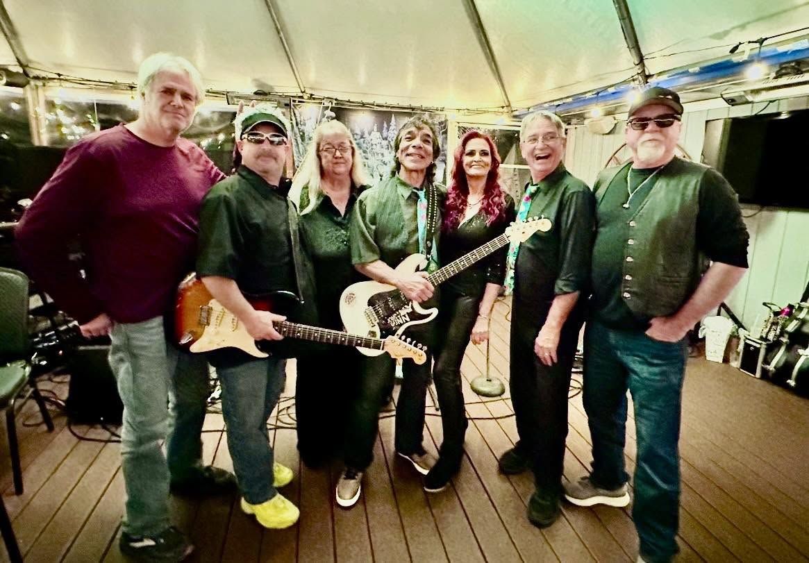 The seven members of The Penetrators Groove Band posing on a deck with their instruments